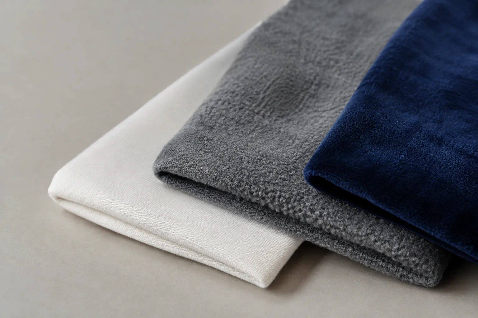 Drie gevouwen stofstalen: glad wit jersey, grijs met een grove lusstructuur en donkerblauw met een fluweelachtig oppervlak